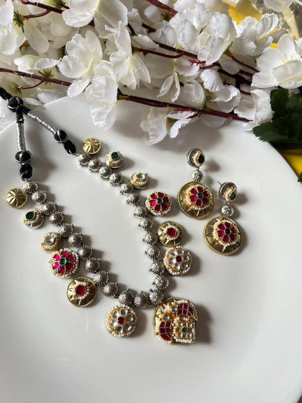 Silver necklace with colorful gemstones and matching earrings on a white plate with flowers in the background